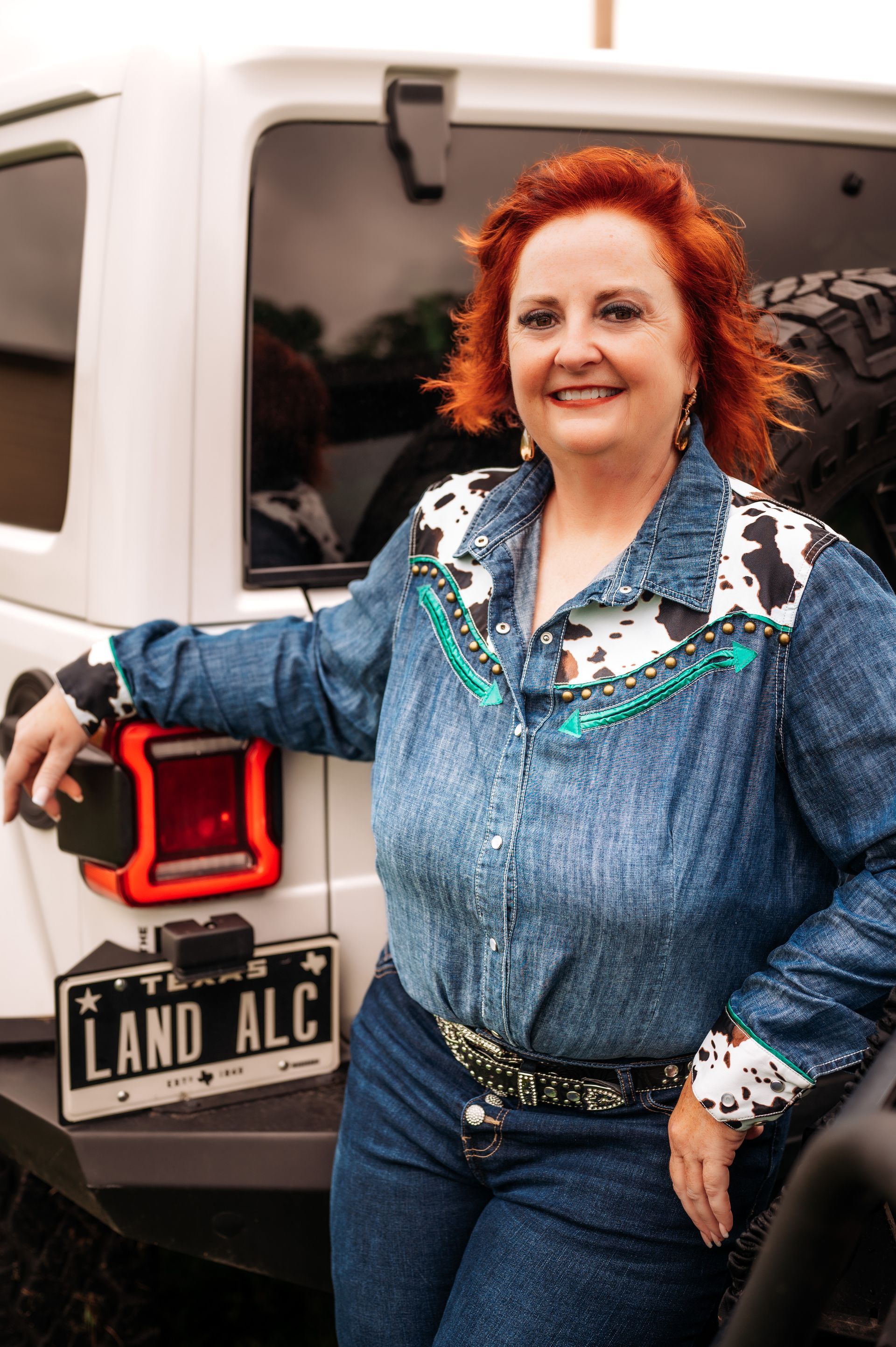 Woman with red hair, smiling, wearing a Western-style denim shirt, leaning on a white Jeep.