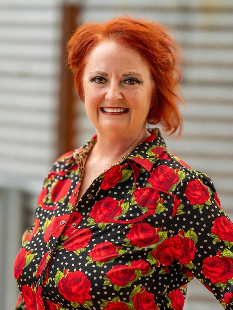 Woman with red hair smiles, wearing a black shirt with red rose pattern, standing outdoors.