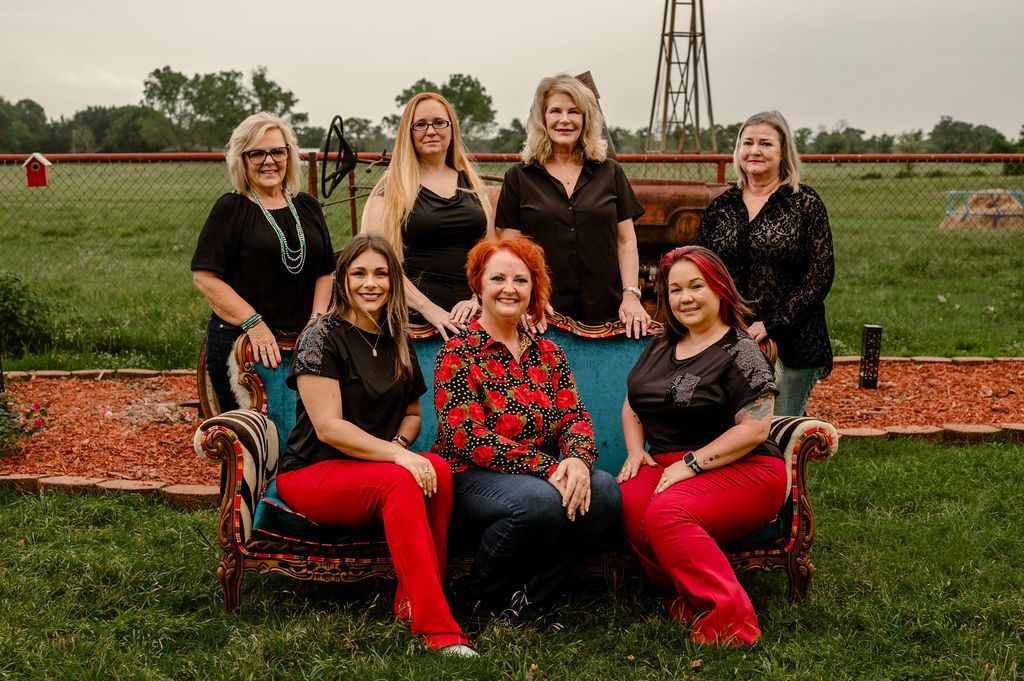 Group of seven women posing on a teal sofa in a grassy field. Most wearing black tops and red pants.