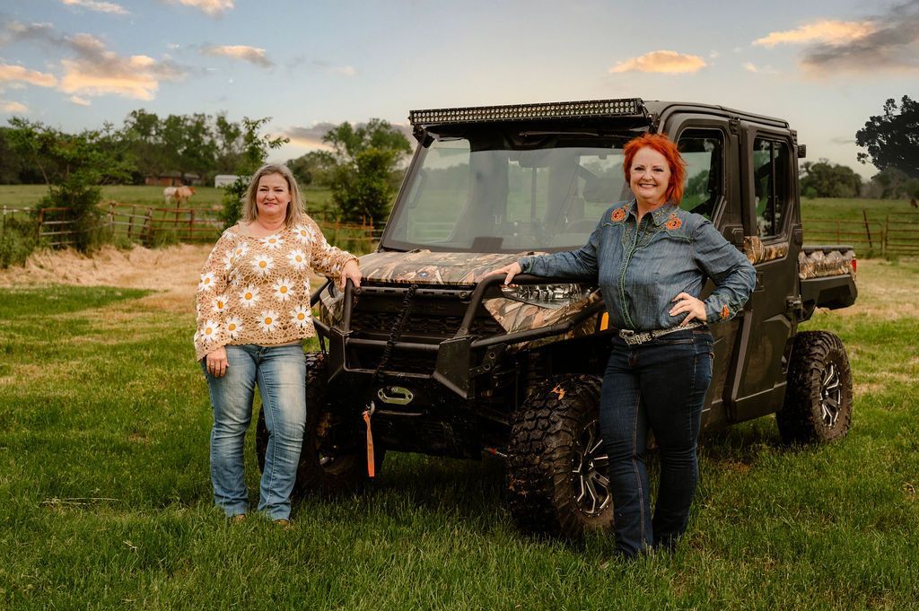 Two women in jeans stand beside a camouflage UTV in a grassy field at sunset.
