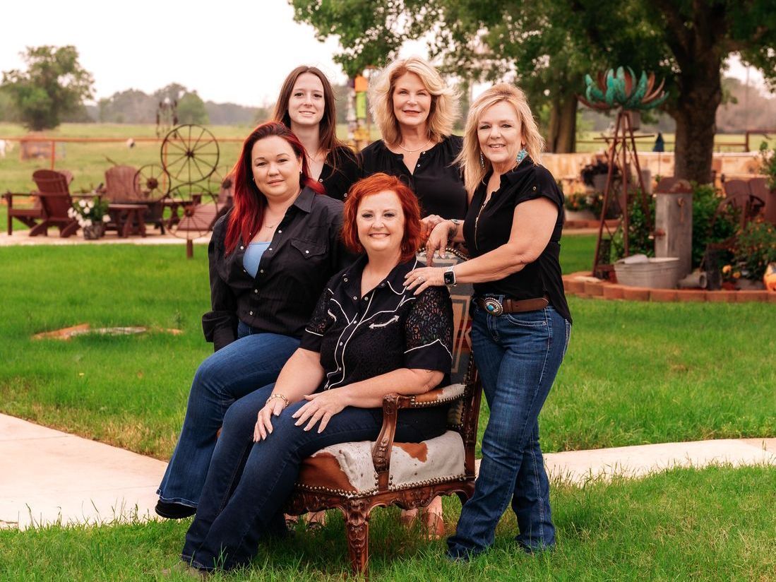 Five women posing outdoors in a grassy area; some seated. All are wearing jeans and black tops.