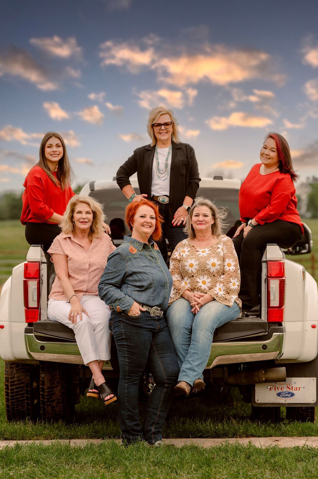 Six women posing on a truck's tailgate in field at sunset. Smiling, wearing casual clothes.