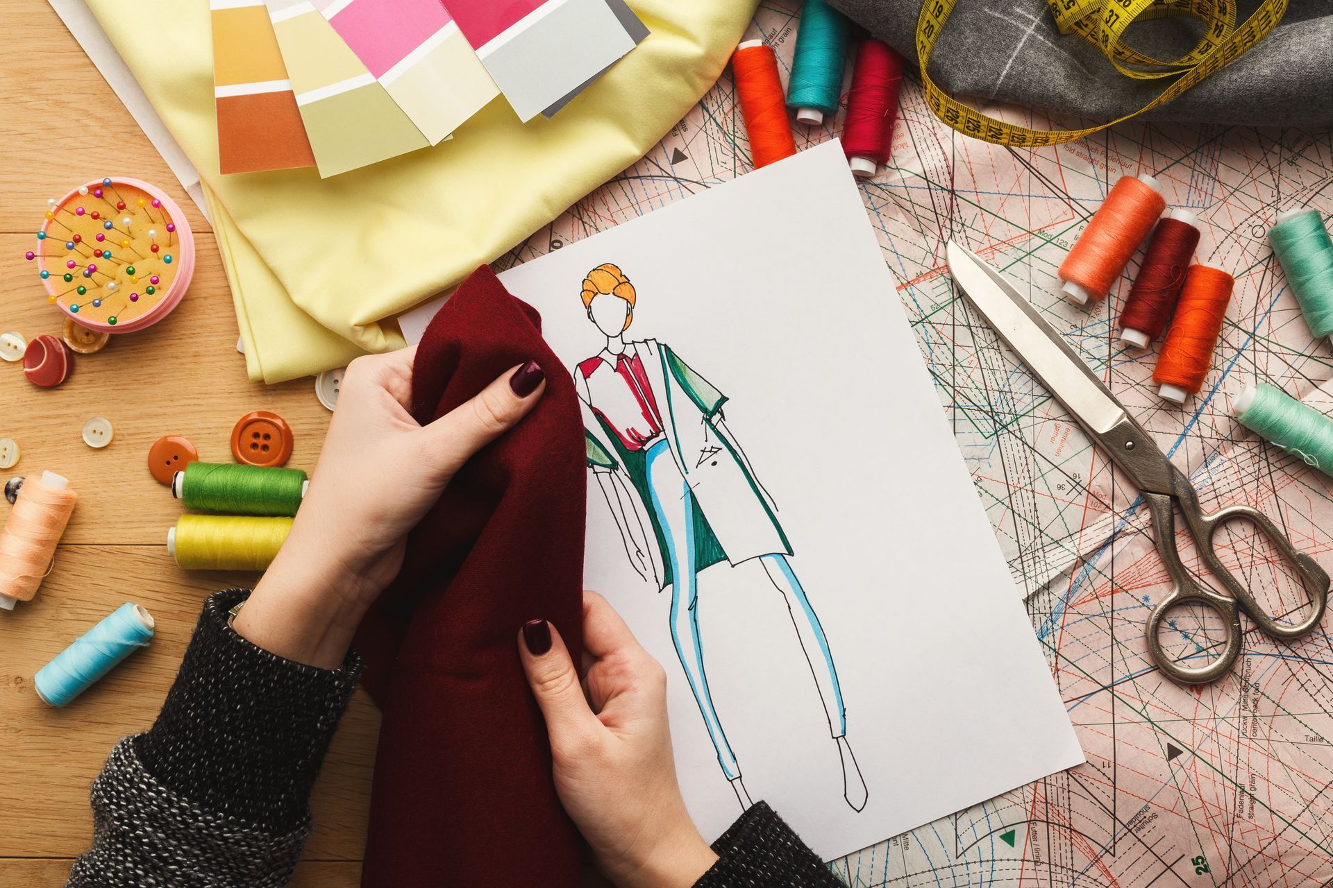 A fashion designer holds a maroon fabric swatch over a sketch, surrounded by thread spools, scissors, and a pattern sheet.