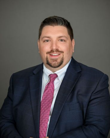 Professional portrait of a man in a navy suit and pink tie against a gray background