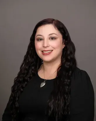 Portrait of a smiling person with long dark hair wearing a black top and necklace on a gray background