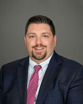 Professional headshot of a man in a navy suit, light blue shirt, and pink tie on a gray background