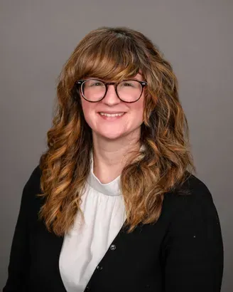 Smiling person with wavy brown hair and glasses, wearing a black blazer and white blouse on a gray background