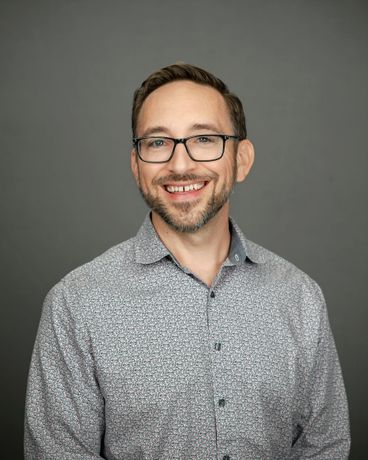 Smiling man in glasses and a patterned button-up shirt against a gray background