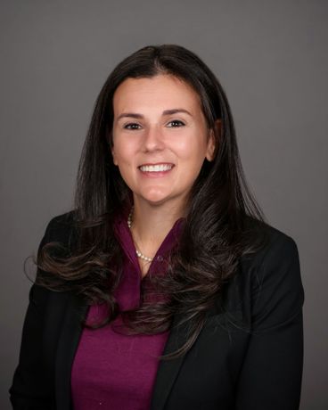 Studio headshot of a smiling woman in a black blazer and purple top against a gray background