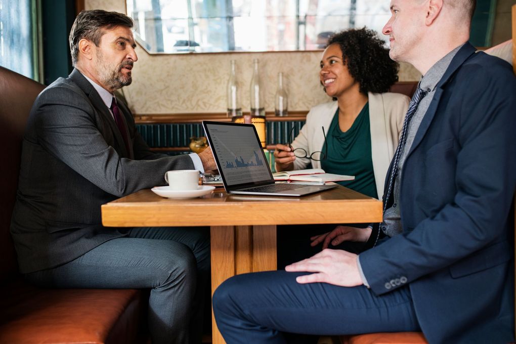 Three people collaborate around a table with laptops near large windows.