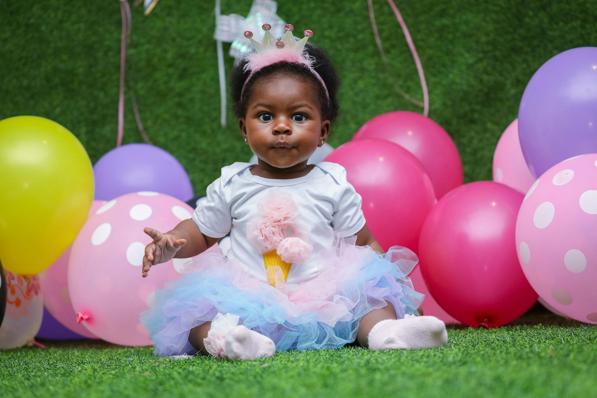 A baby girl is sitting on the grass in front of balloons.