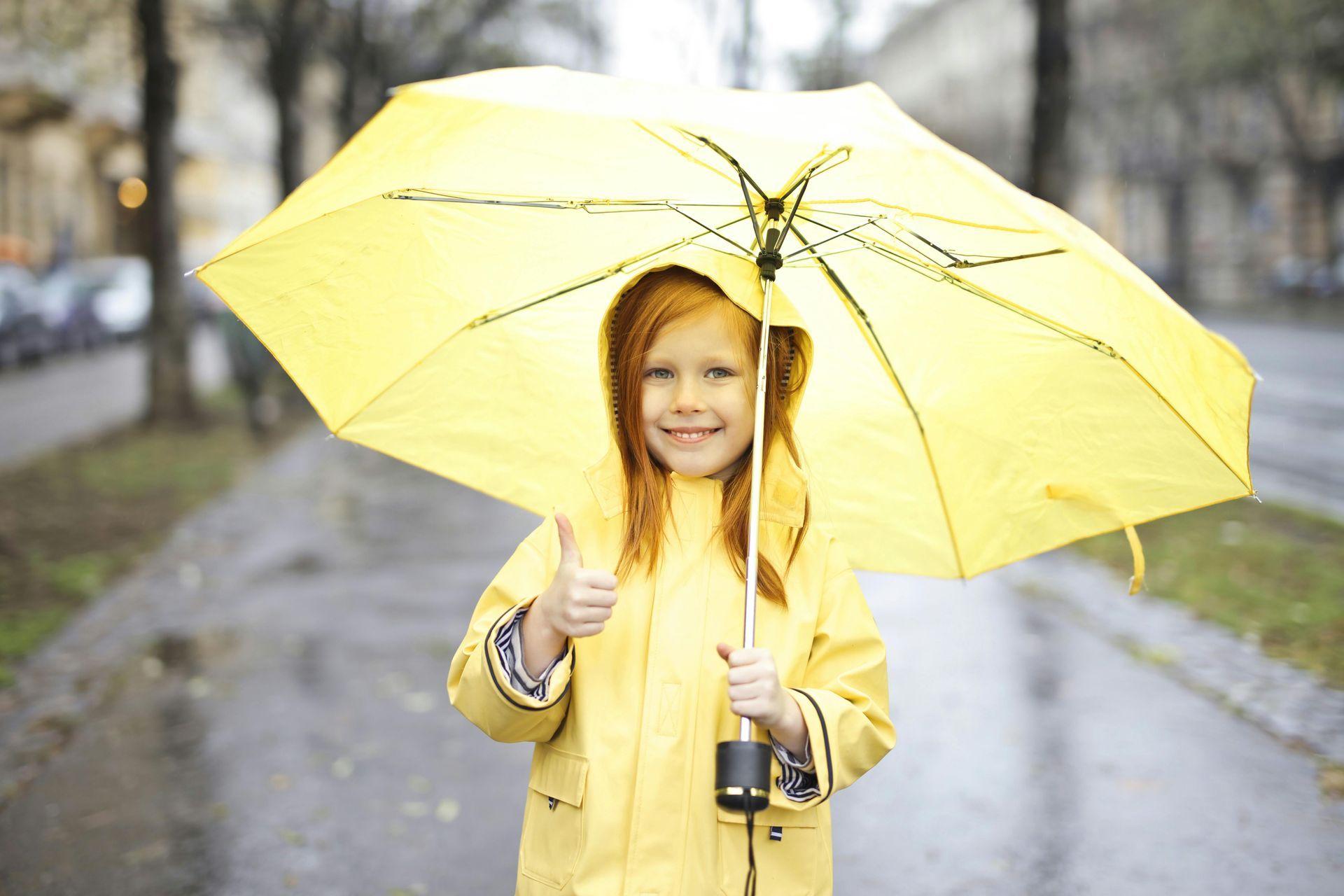 A little girl in a yellow raincoat is holding a yellow umbrella and giving a thumbs up.
