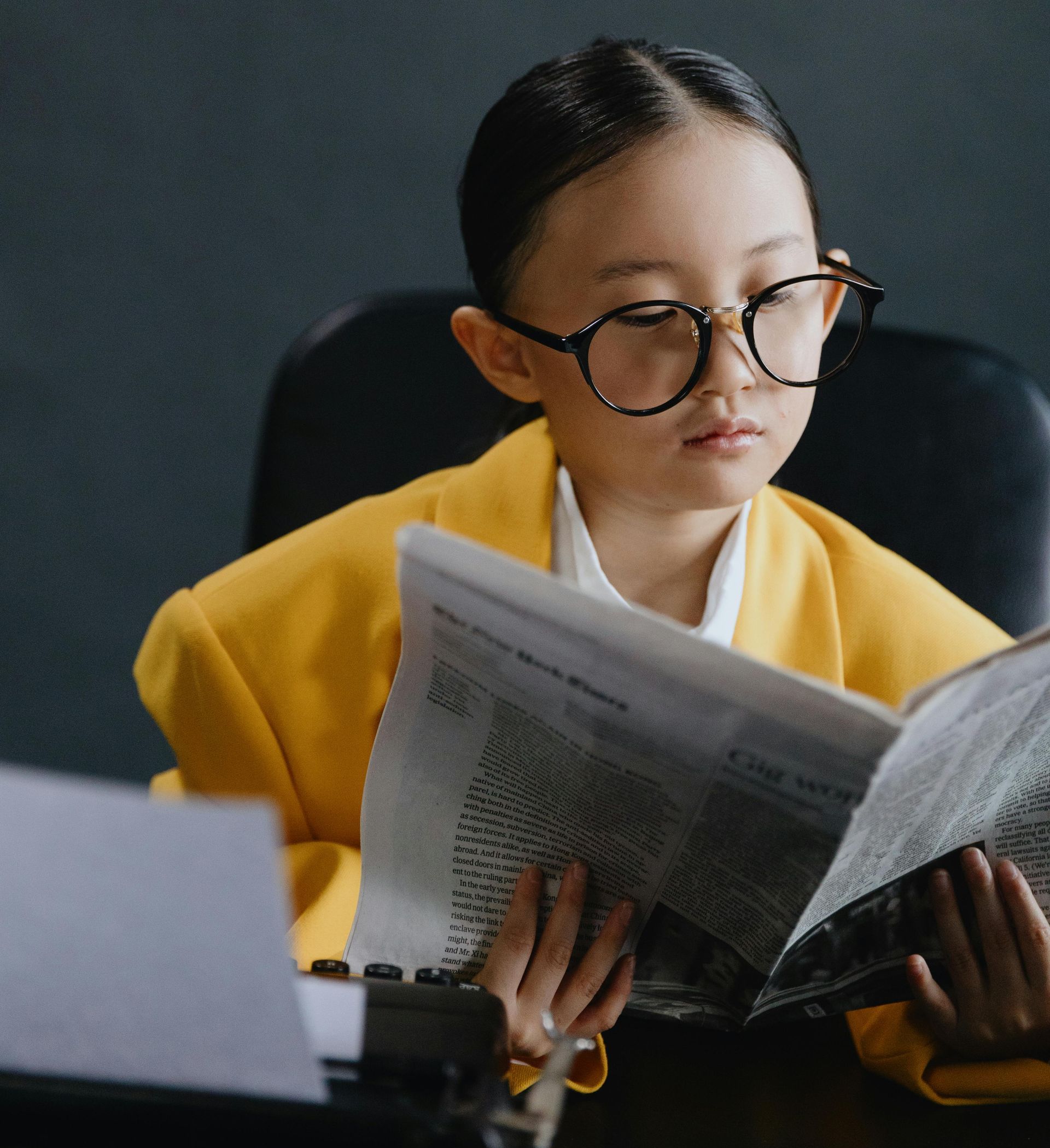 A young girl wearing glasses is reading a newspaper