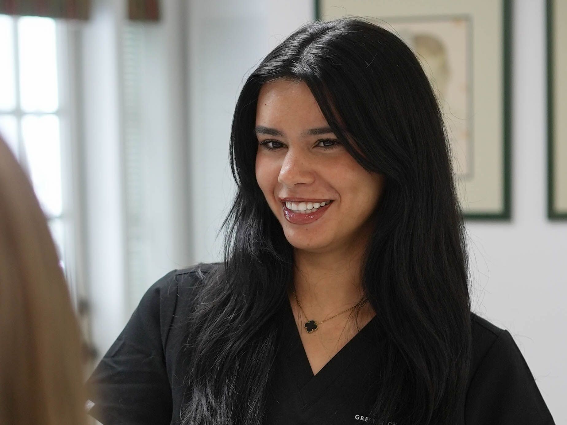 A woman with long black hair is wearing a black scrub top and smiling