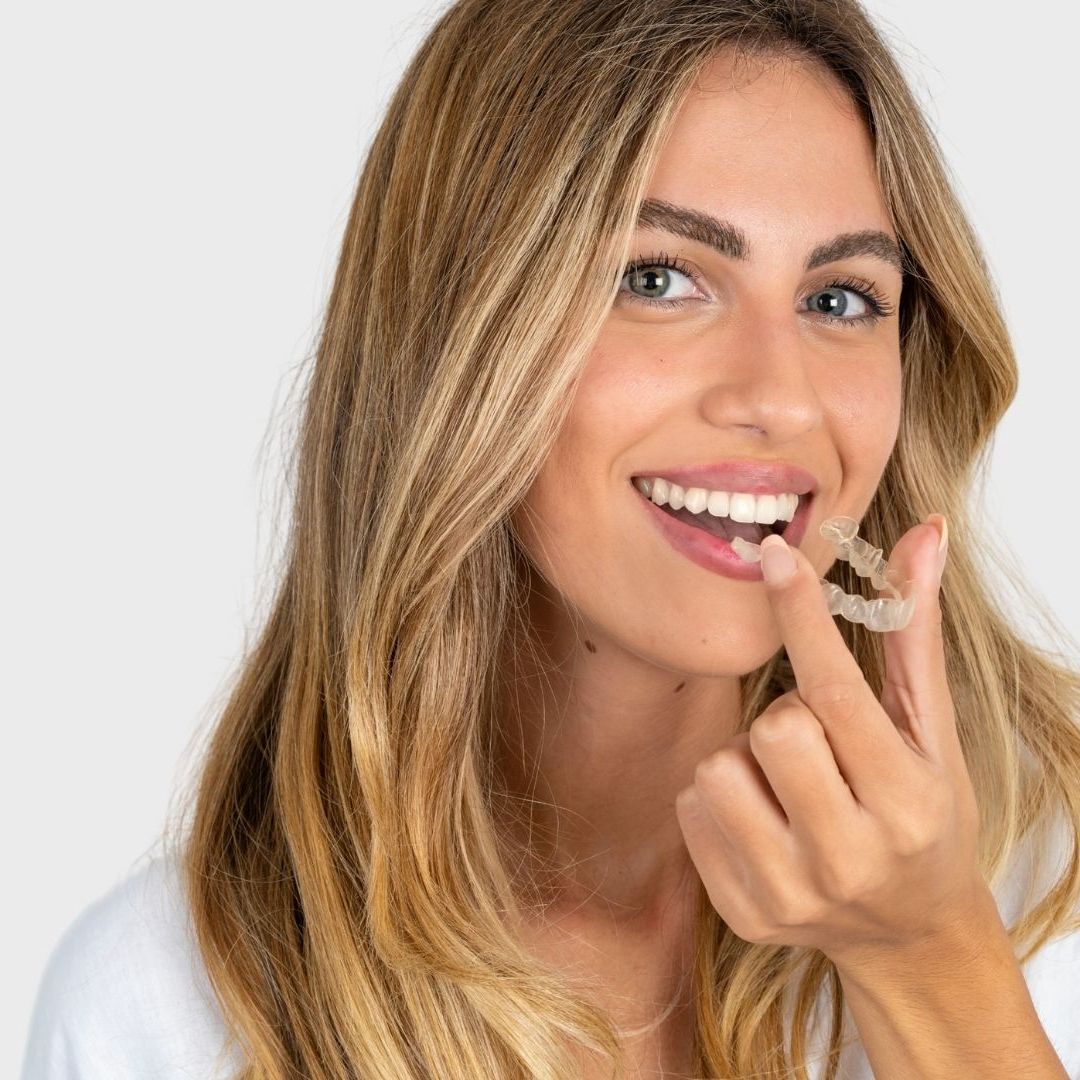 A woman with long blonde hair is smiling and holding a clear braces in her hand