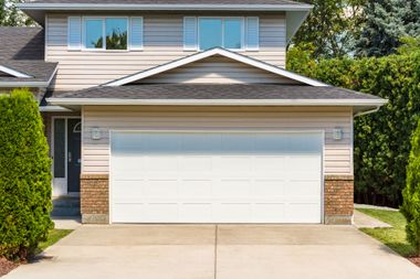 A beige two-story suburban house with a large white double garage door and a front entrance, framed by green shrubs.