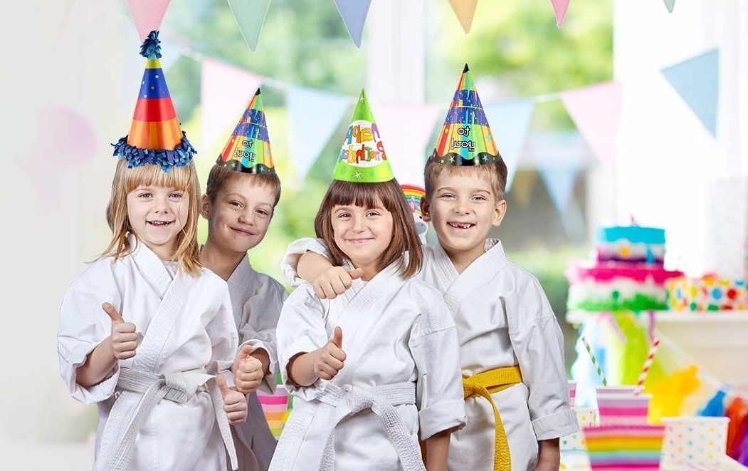 A group of children wearing karate uniforms and party hats are giving a thumbs up.