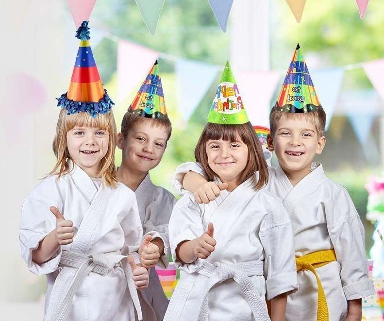 A group of children wearing karate uniforms and party hats are giving a thumbs up.