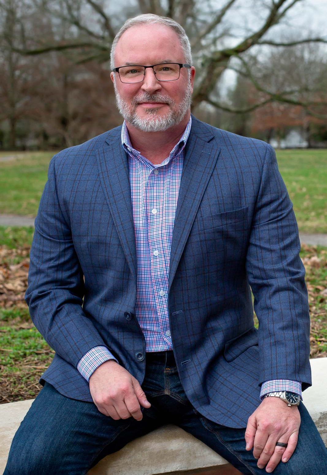Man in glasses and blue blazer, hands in pockets, stands in front of a brick wall.