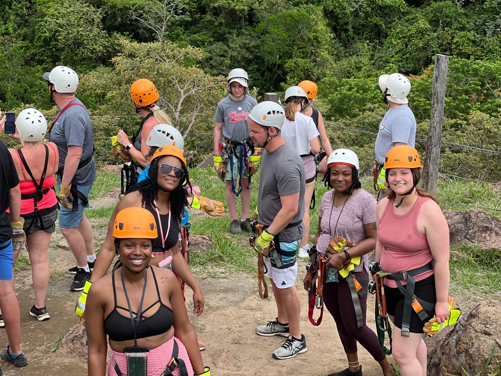 A group of people wearing helmets and climbing harnesses standing on a dirt path in a wooded area.