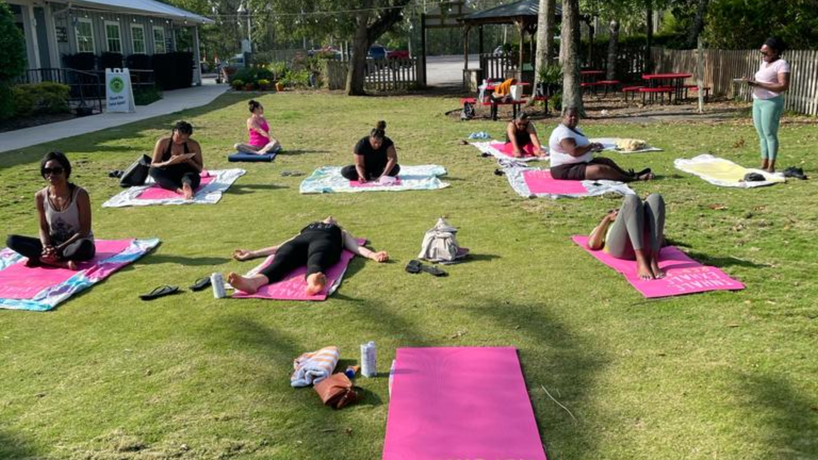 A group practices yoga on colorful mats in a sunny, grassy park, with an instructor standing nearby.