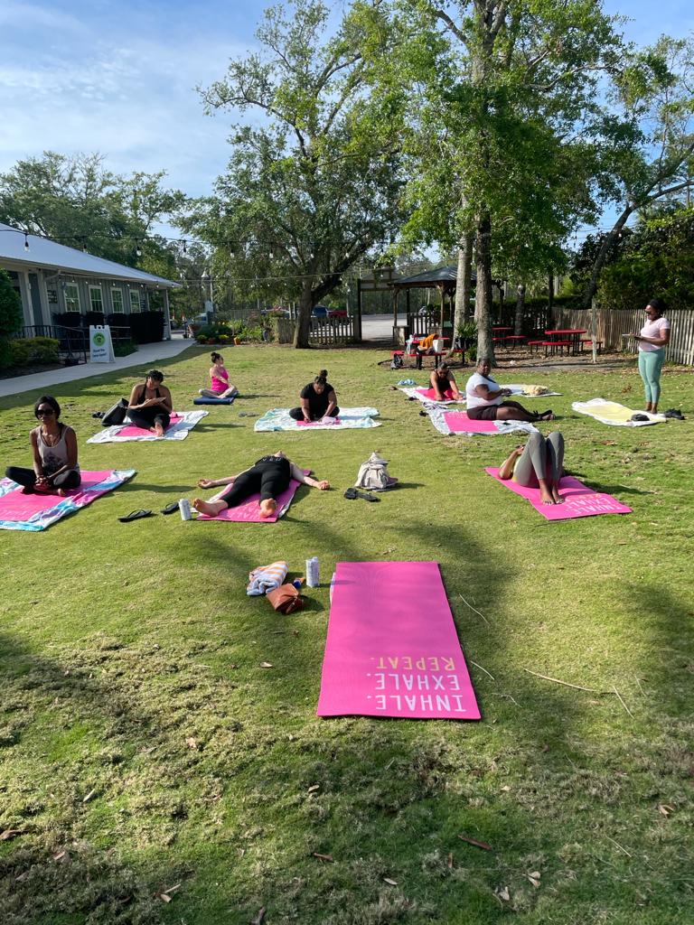 People practicing yoga on colorful mats in a sunny outdoor grassy area near a building and trees.
