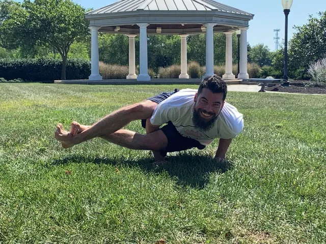A person with a beard smiling while performing an arm-balance yoga pose on a grassy lawn in front of a gazebo.