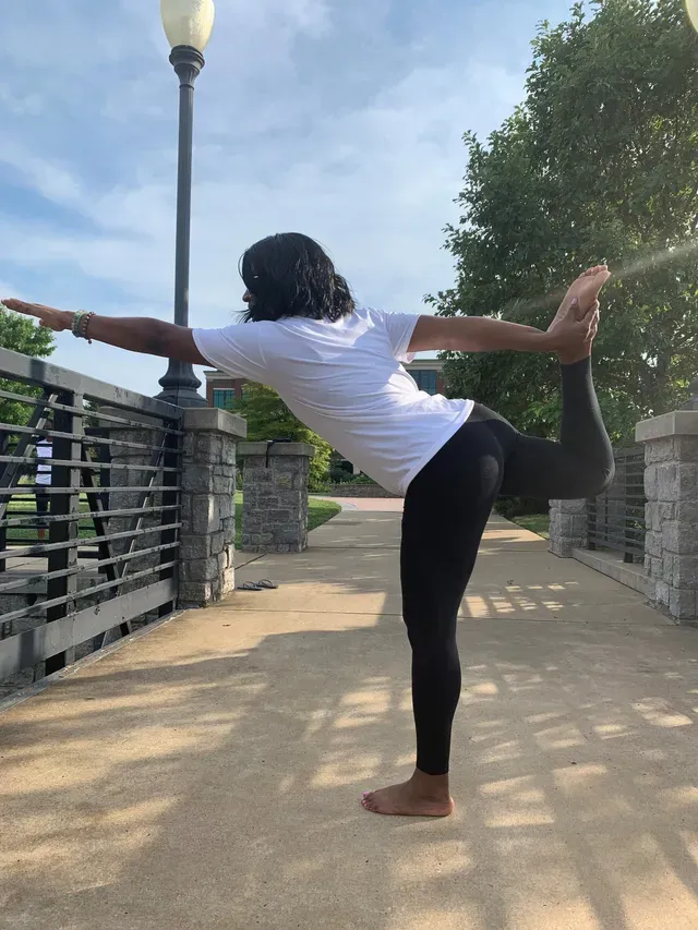 A person in a white shirt and black leggings performs a yoga dancer pose on a stone path next to a bridge railing.