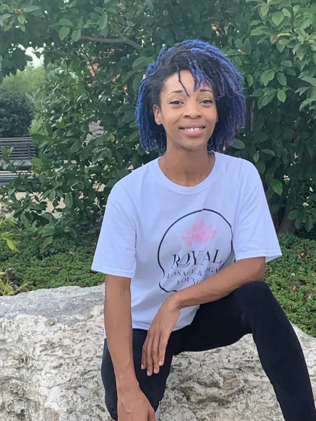 A person with vibrant blue hair wearing a white t-shirt, kneeling on a rock in front of a leafy green backdrop.
