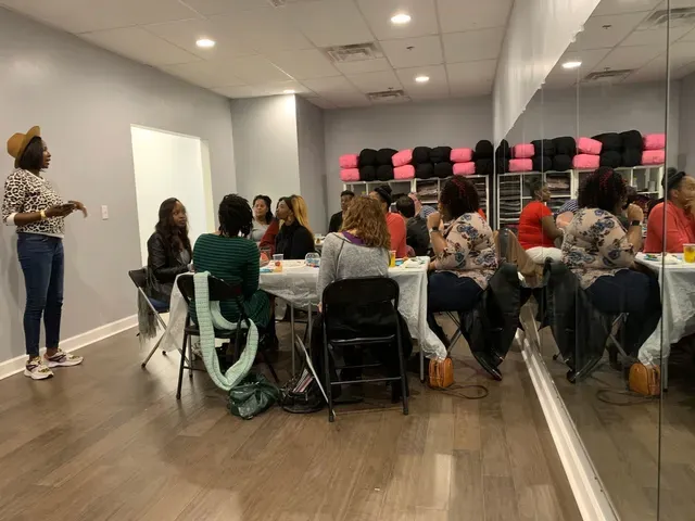 A woman stands and speaks to a group of people seated around a table at a studio meeting, with mirrors lining one wall.