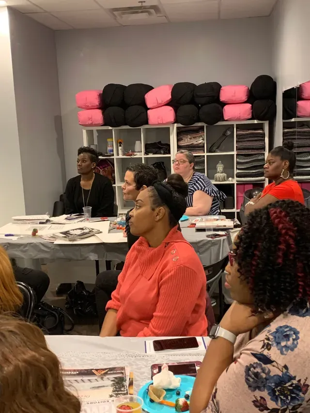 A group of people sits at tables in a studio, listening to a presentation with yoga mats and blocks on shelves behind them.