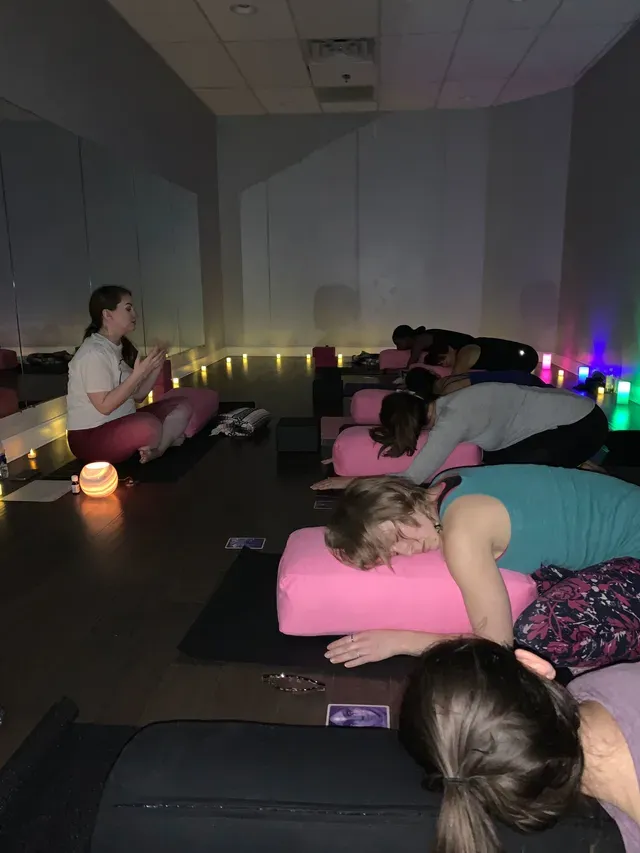 A group of people in a dim yoga studio practice restorative poses while lying on pink bolsters with an instructor nearby.