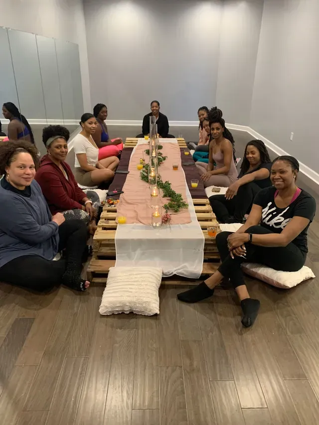 A group of people seated on cushions around a low, pallet-style dining table in a studio with wooden floors.