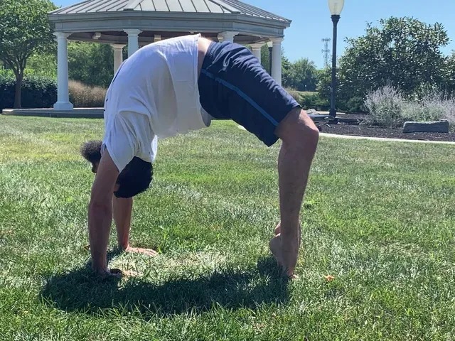 A person performs a wheel pose yoga stretch on a grassy lawn in front of a gazebo on a sunny day.