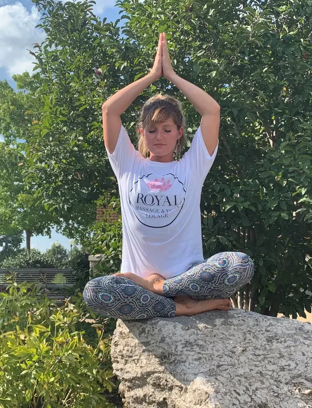 A person in a white t-shirt and patterned leggings meditates in a cross-legged pose on a large rock outdoors.