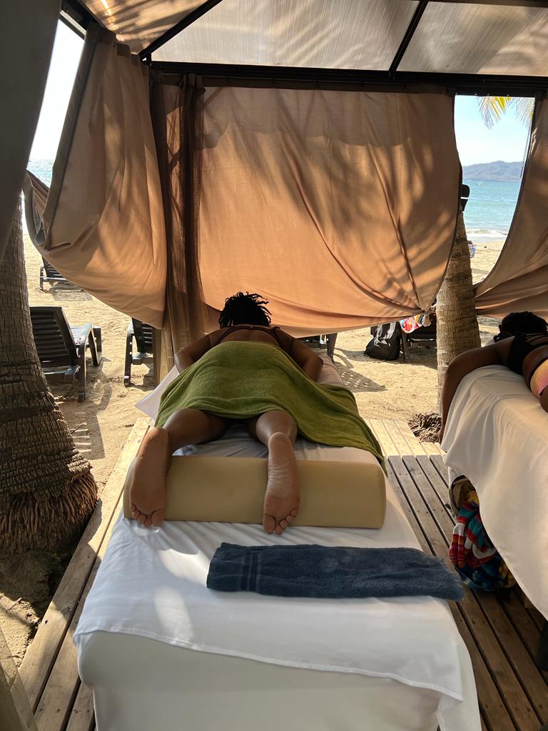 A person lies face down on a massage table under a beach cabana with a green sarong, near the ocean.