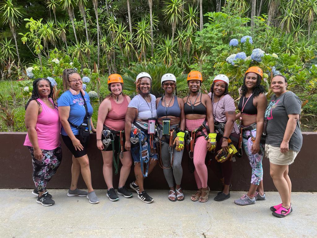 A group of nine people wearing climbing gear and helmets pose together in front of a hedge with light blue flowers.