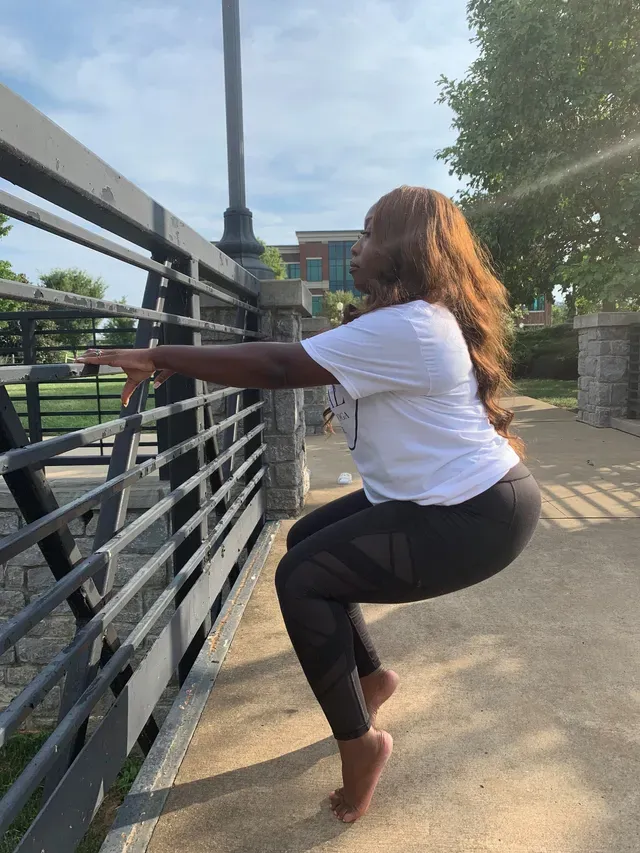 A person in a white t-shirt and black leggings performing a barefoot squat while holding onto a metal railing outdoors.