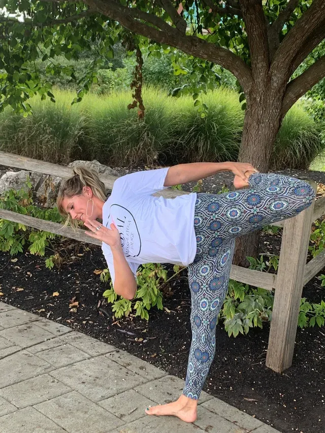 A person in a white shirt and patterned leggings performs a balancing dancer yoga pose outdoors near a wooden fence.