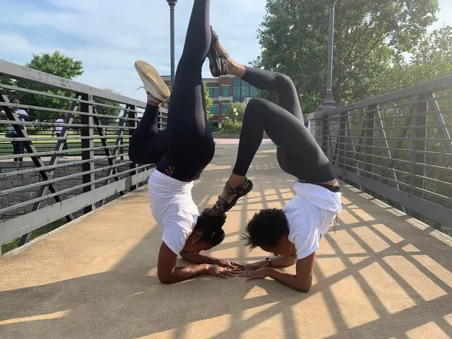 Two people in white shirts and black leggings perform a synchronized forearm stand, connecting their feet on a bridge.