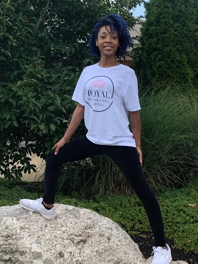 A person with blue hair wearing a white T-shirt and black leggings stands with one foot on a large rock outdoors.