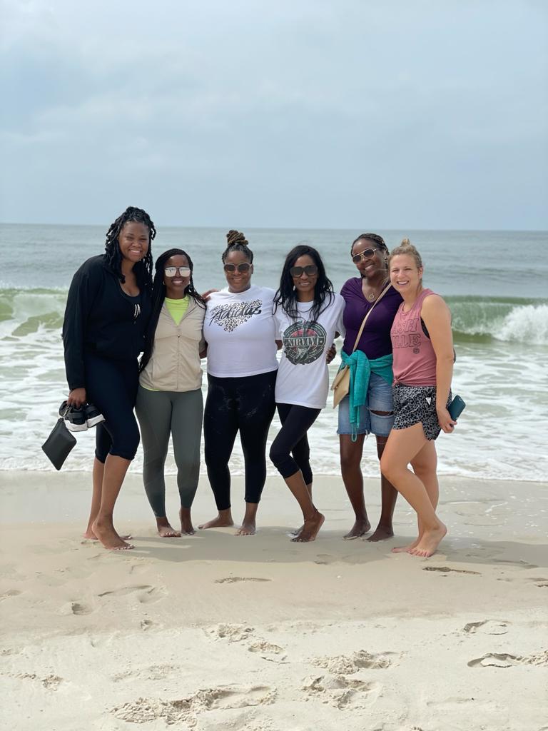 Six people stand in a line on a sandy beach by the ocean, posing together and smiling at the camera on an overcast day.