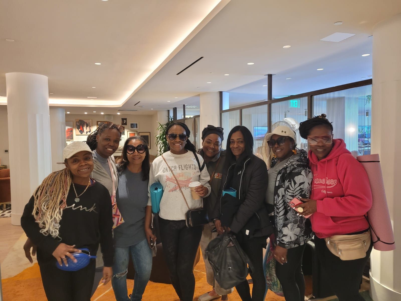 A group of eight people smiling while standing in a hotel lobby, some holding bags and wearing athletic clothing.