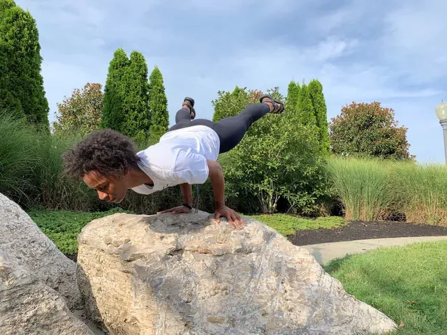 A person performing a yoga arm balance pose on a large rock in an outdoor park setting with green trees and a blue sky.