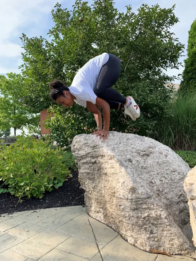 A person in athletic wear performs a crow pose yoga balance on top of a large boulder in a garden setting.