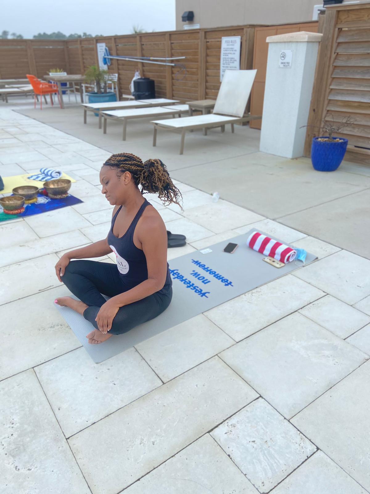 A person sits in a meditative pose on a yoga mat outdoors on a tiled pool deck, with lounge chairs in the background.