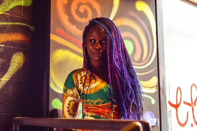 A person with long, vibrant purple braided hair wearing a patterned top stands before a colorful abstract mural.