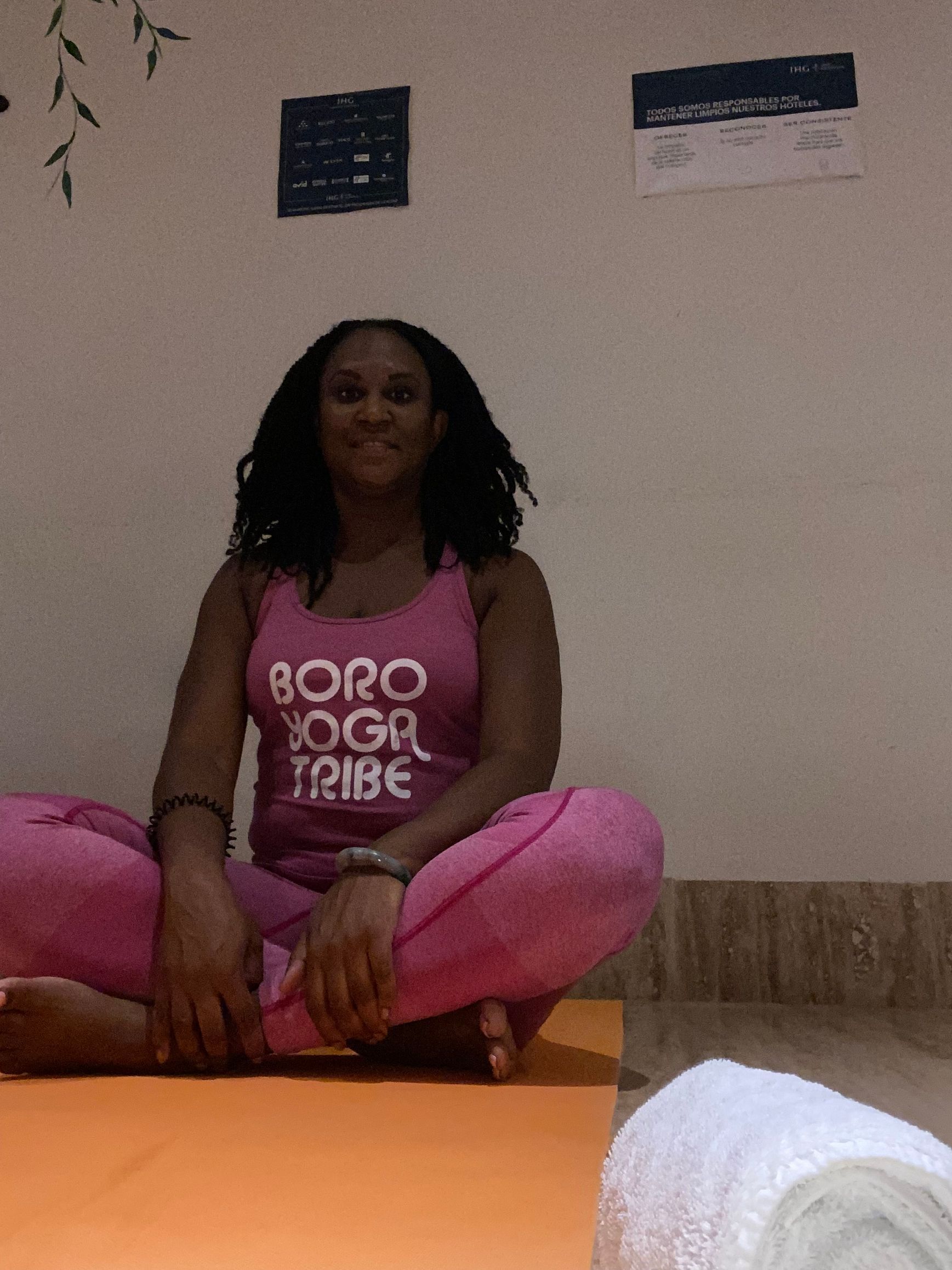 A person with dark hair sits in a cross-legged yoga pose on an orange mat, wearing a pink tank top and matching leggings.