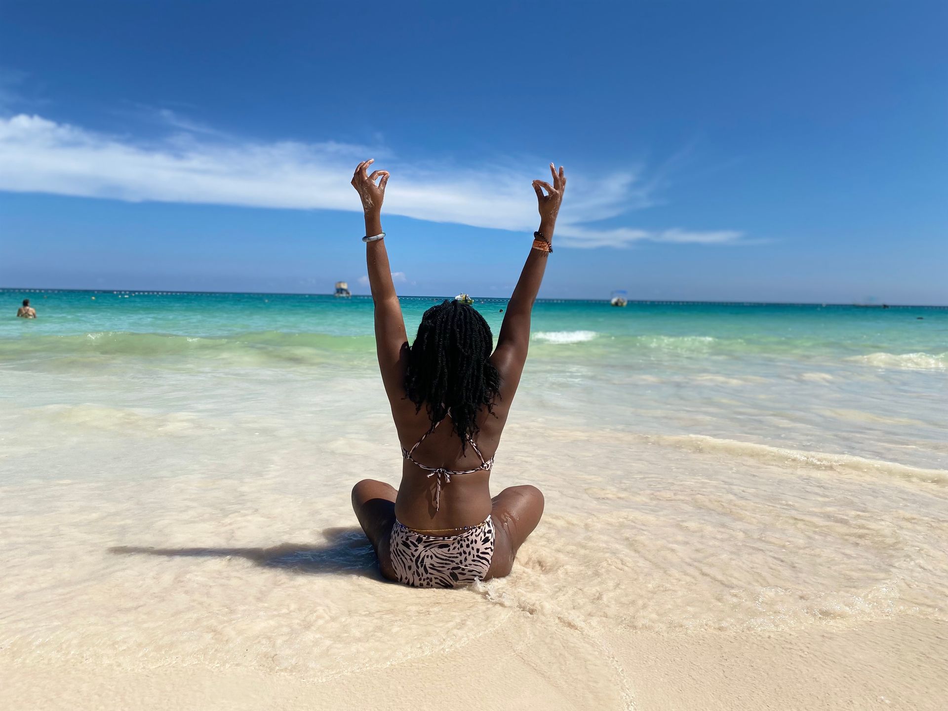 A person sits cross-legged on a sunny beach with arms raised in a yoga pose, facing the turquoise ocean under a blue sky.