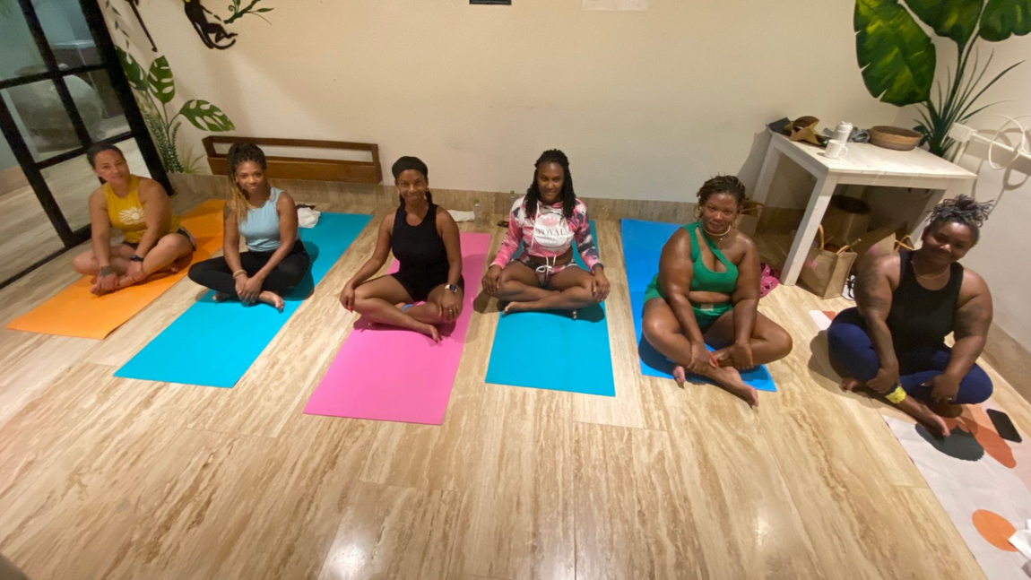 Six people sit cross-legged on colorful yoga mats in a bright studio, facing forward.